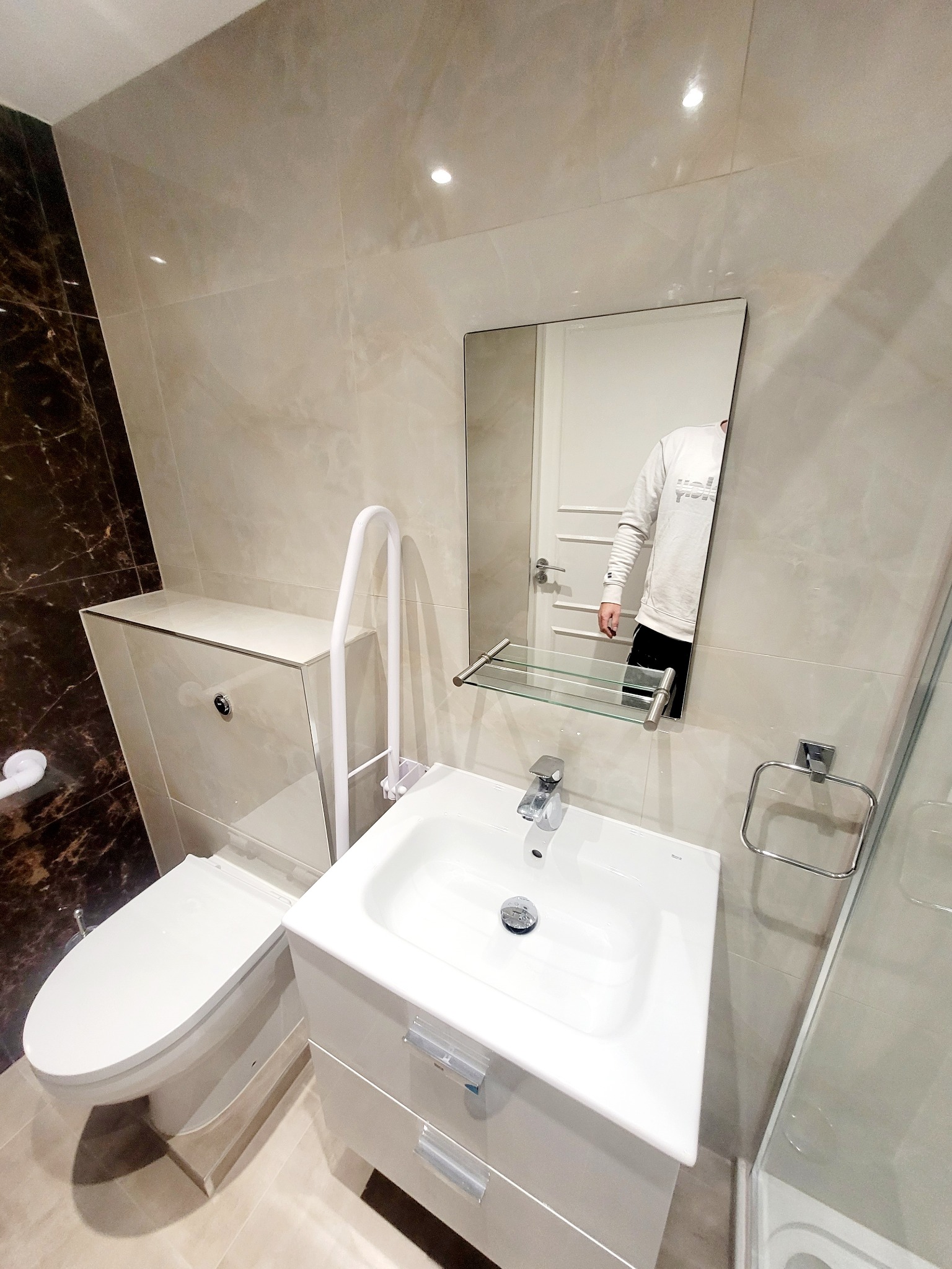 Nowoczesna, jasna łazienka z umywalką na szafce, toaletą z ukrytą spłuczką i uchwytem dla osób z niepełnosprawnościami, wykończona płytkami imitującymi marmur, z odbiciem osoby w lustrze.