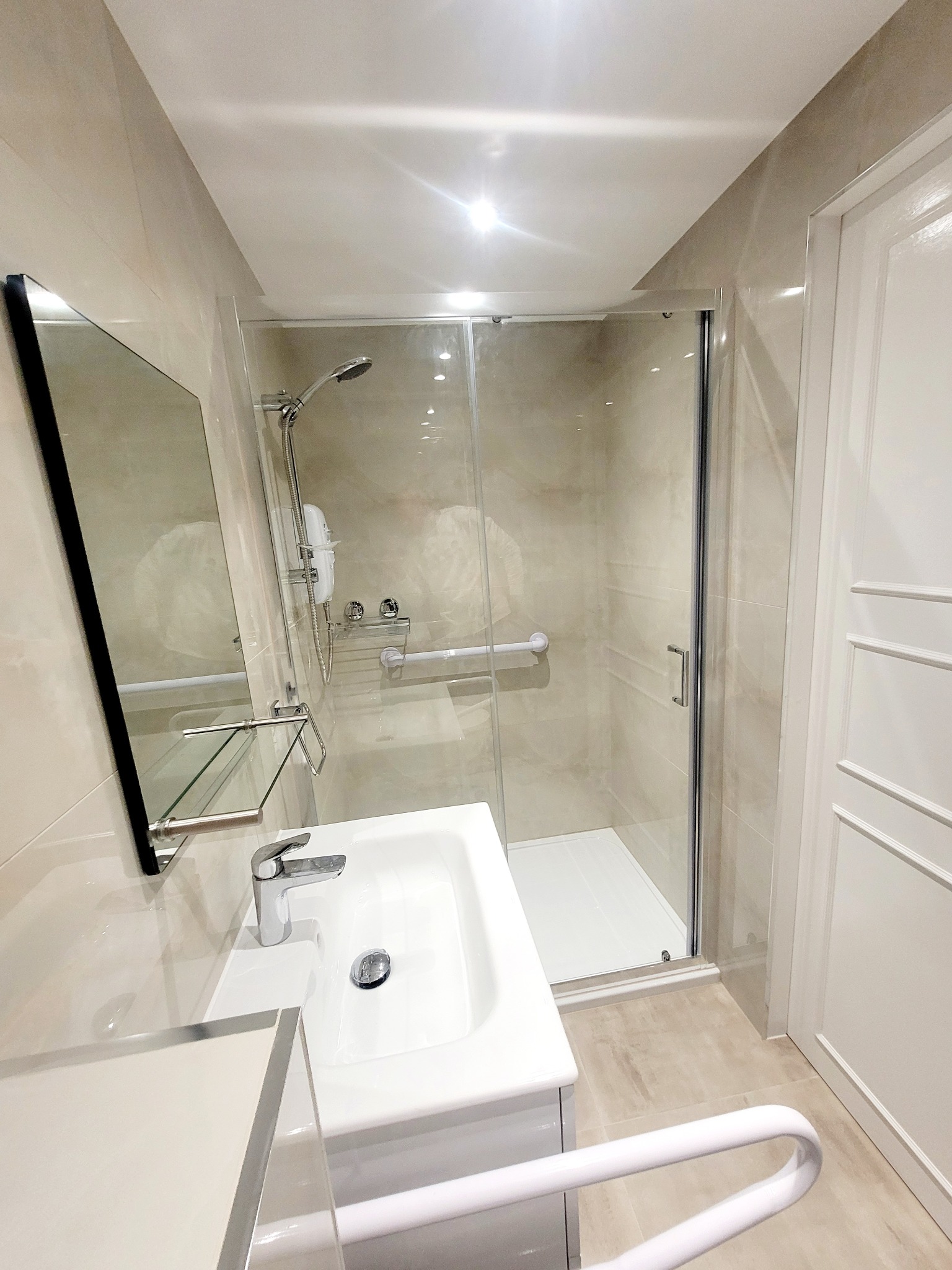 Jasna łazienka z beżowymi płytkami, kabiną prysznicową ze szklanymi drzwiami i uchwytem dla niepełnosprawnych, umywalką nablatową i lustrem w czarnej ramie.