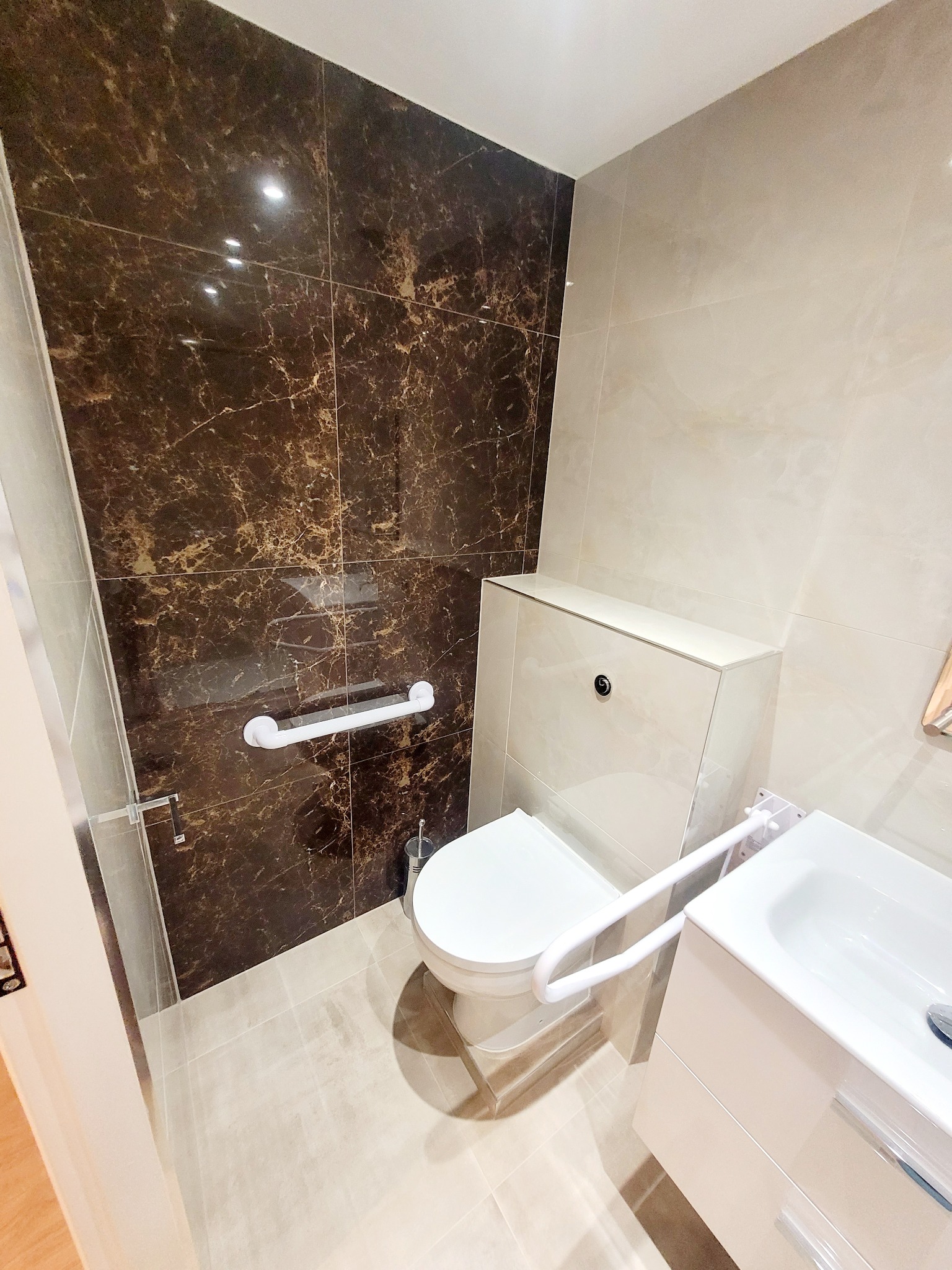 Wykończona łazienka z brązową marmurową ścianą, toaletą podwieszaną i umywalką z szafką, wyposażona w uchwyty dla osób niepełnosprawnych.