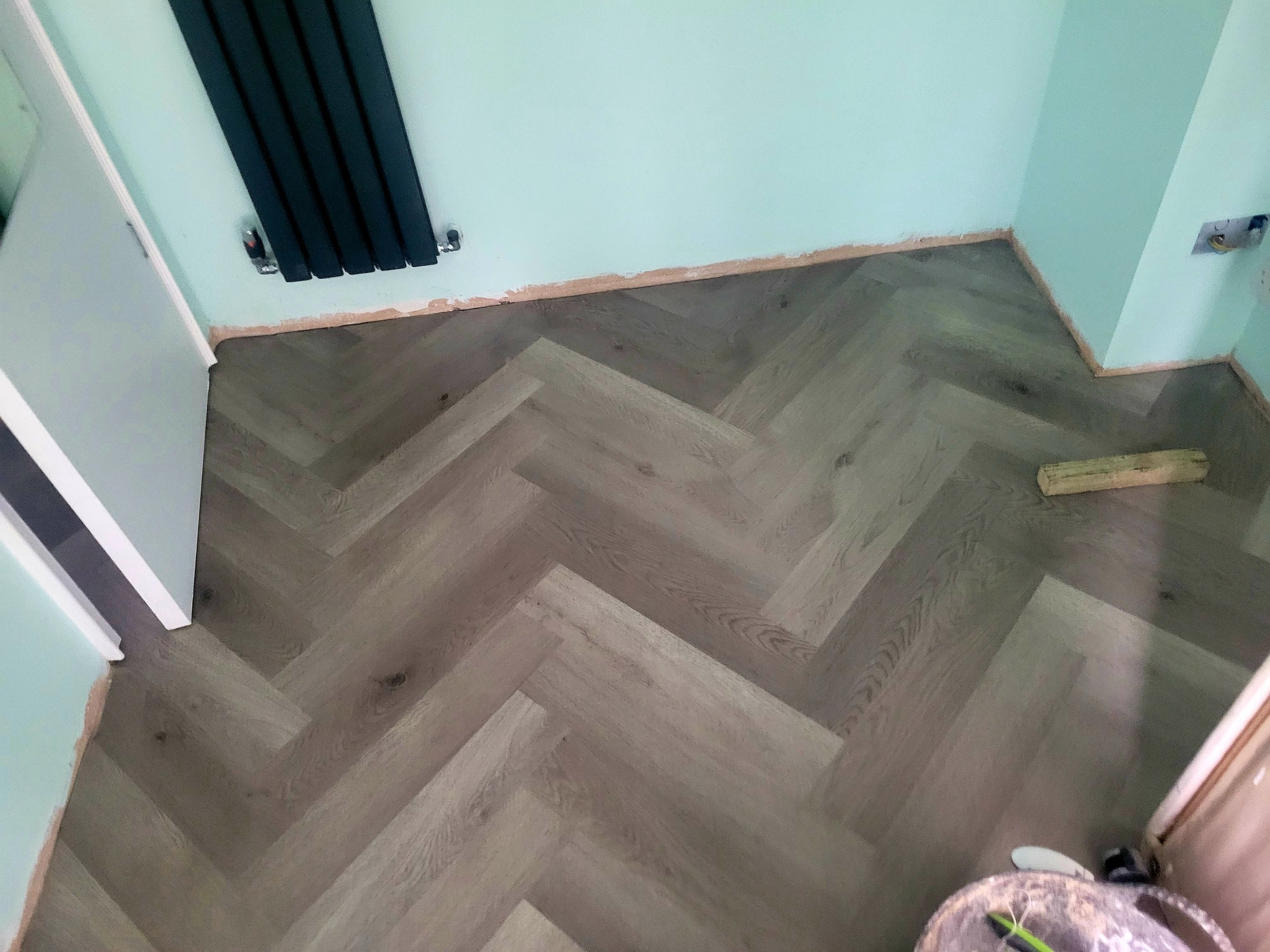 Ułożona pod kątem jodełki podłoga z paneli w odcieniach szarości, widoczny fragment ściany pomalowanej na miętowo, czarny grzejnik oraz niezakończone prace wykończeniowe przy ścianie.