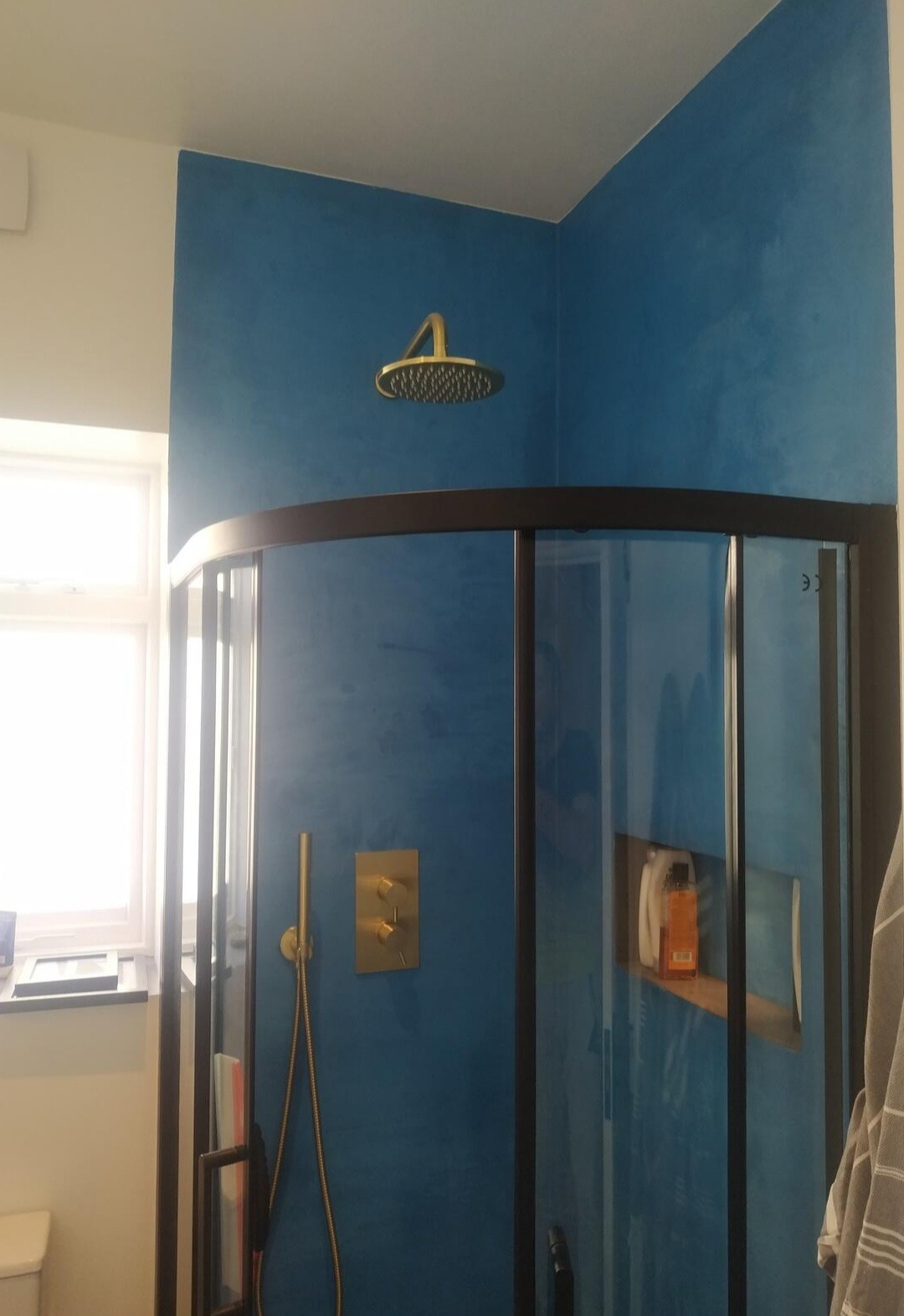 Niebieska łazienka z kabiną prysznicową o czarnych profilach, złotą armaturą i ścianami wykończonymi gładzią imitującą efekt betonu.