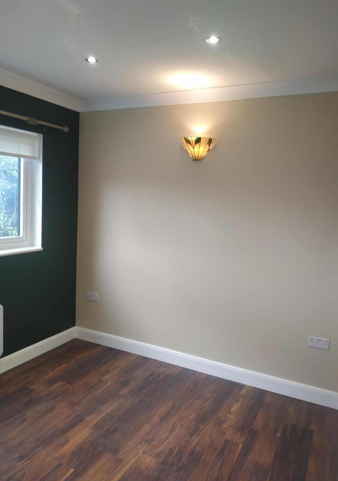 Pokój po remoncie z ciemnozieloną ścianą, panelami podłogowymi w odcieniach brązu i beżową ścianą z ozdobnym kinkietem.