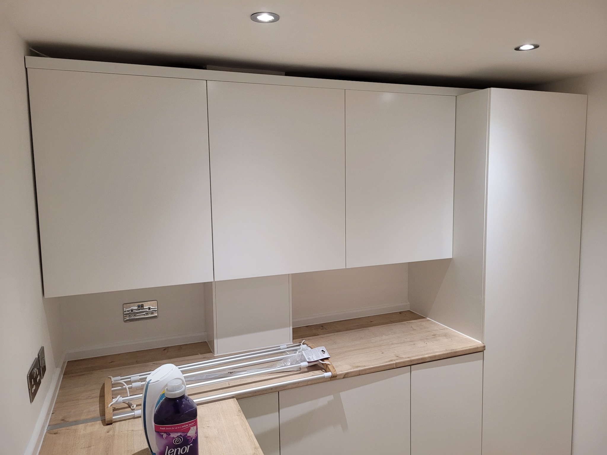 Białe szafki kuchenne z drewnianym blatem, widoczny nowy rozkładany wieszak na pranie i płyn do płukania Lenor, gniazdka elektryczne na ścianie.