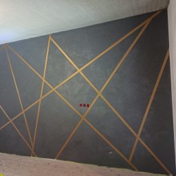 Pro-Bud Krzysztof Kargut - Szara ściana z dekoracyjną strukturą i złotymi geometrycznymi pasami, widoczne gniazdka elektryczne.