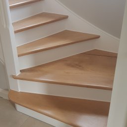 Pro-Bud Krzysztof Kargut - Jasne drewniane schody z białymi podstopnicami i poręczą, widok z dołu, neutralne płytki podłogowe, naturalne światło z okna w tle.