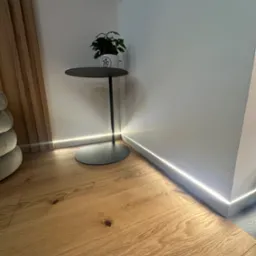 Nowoczesne wnętrze z drewnianą podłogą, listwą przypodłogową z podświetleniem LED, minimalistycznym stolikiem z rośliną w kubku i elementem dekoracyjnym z drewnianymi listwami na ścianie.