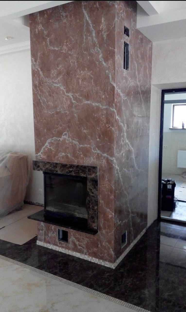 Obudowa kominka z efektem marmuru w odcieniach brązu i bieli, z czarnym obramowaniem paleniska i fragmentem błyszczącej, ciemnej podłogi, widoczne prace wykończeniowe.