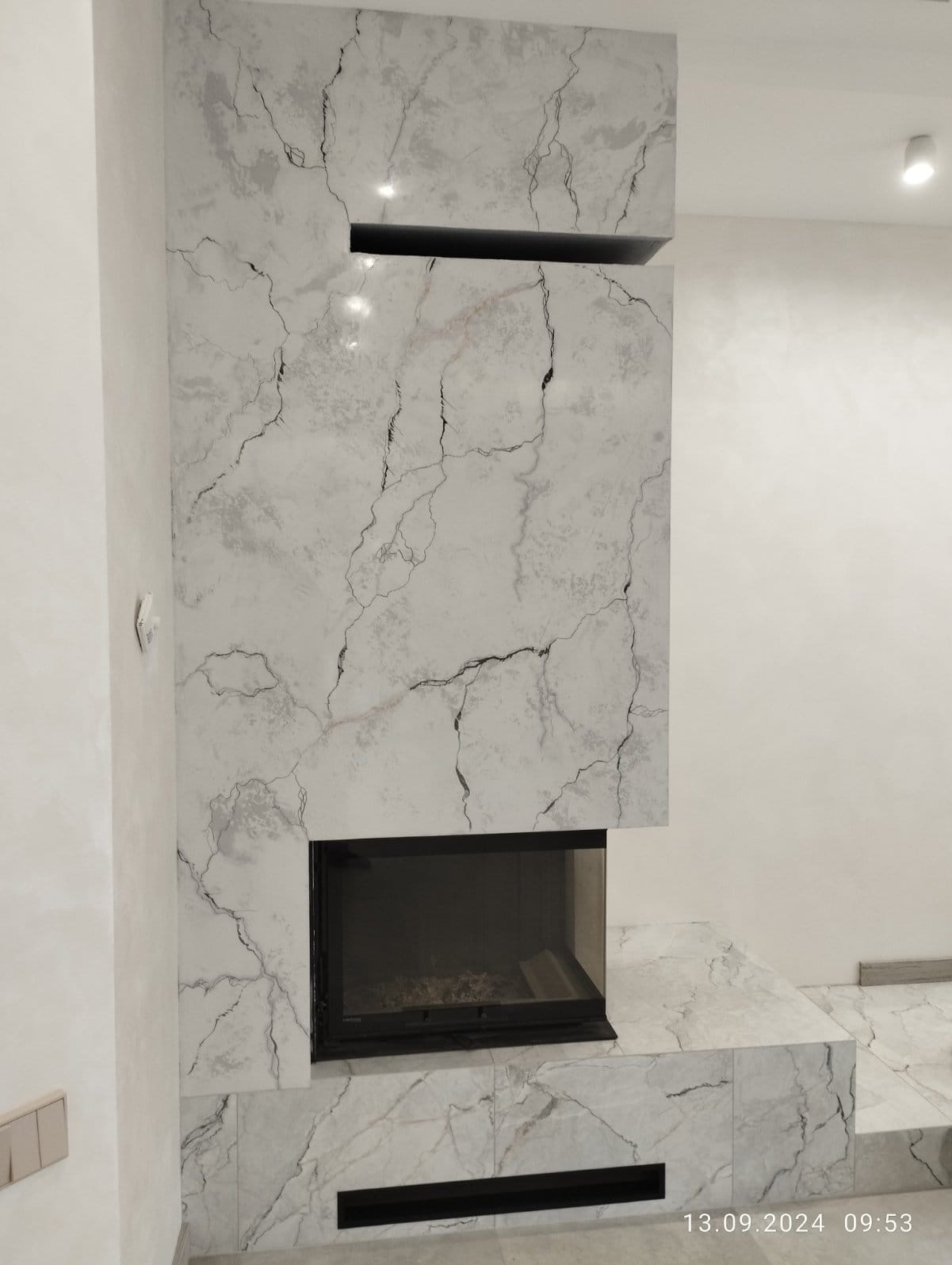 Obudowa kominka z płyt imitujących biały marmur z czarnymi żyłami, z wkładem kominkowym, na tle białej ściany.