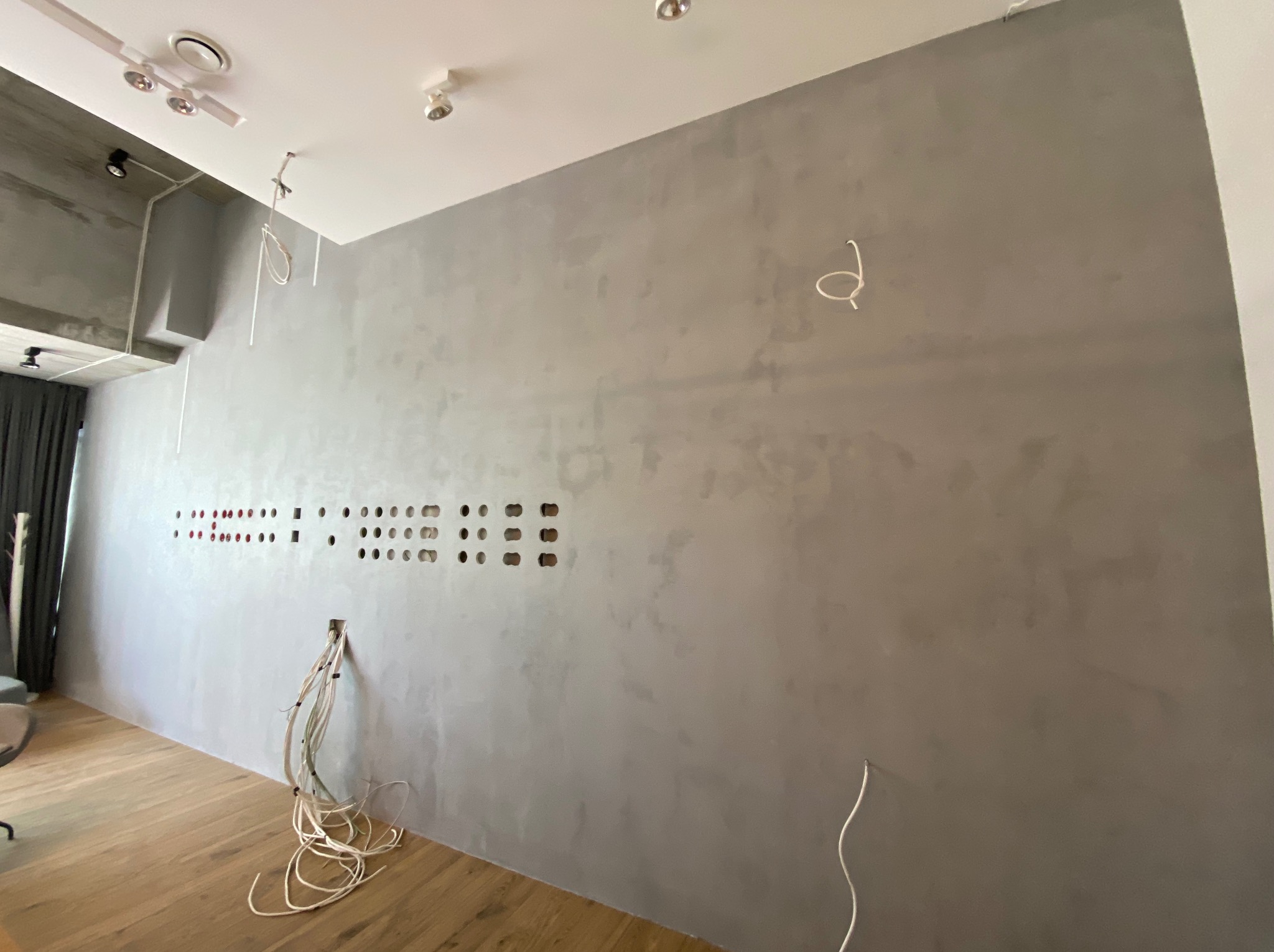 Ściana w szarym odcieniu z widocznymi otworami na przewody elektryczne, przygotowana do montażu instalacji, z luźno zwisającymi kablami.