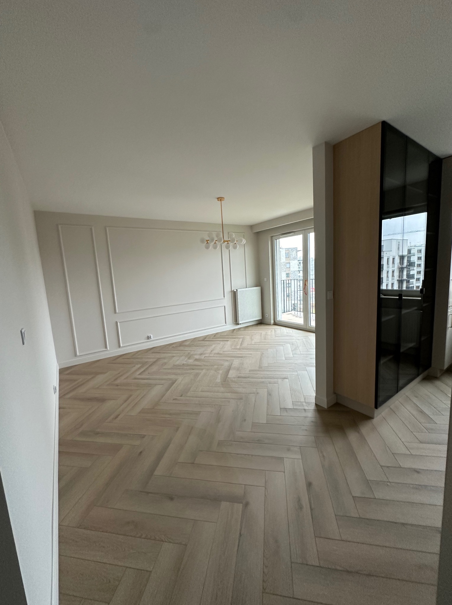 Jasne, przestronne wnętrze po remoncie: parkiet w jodełkę, białe ściany z dekoracyjnymi listwami, złoty żyrandol i czarna szafa w zabudowie.