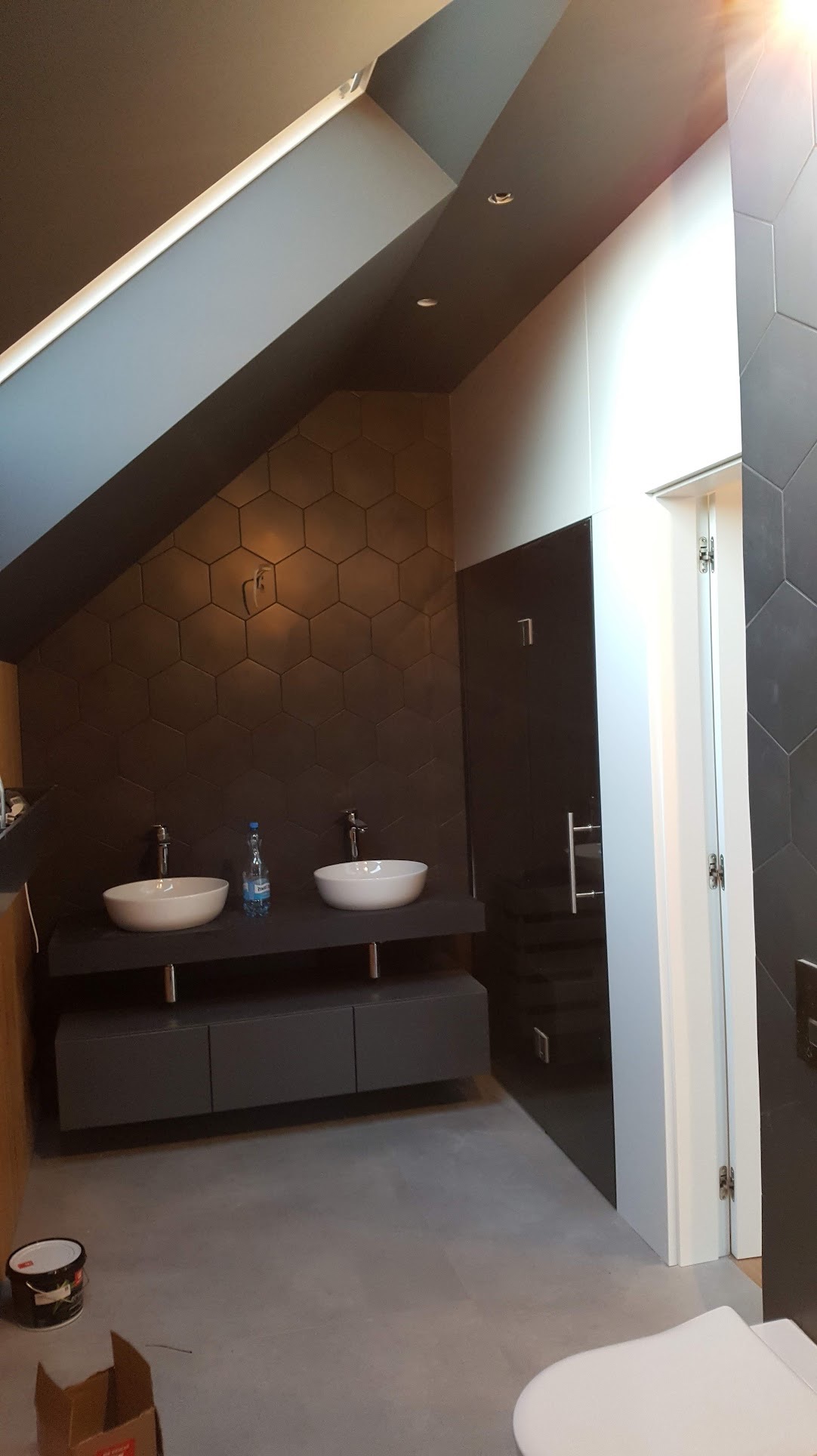 Nowoczesna łazienka na poddaszu z dwoma umywalkami nablatowymi, heksagonalnymi płytkami na ścianie, szarą szafką pod umywalkami i szklanymi drzwiami do kabiny prysznicowej.