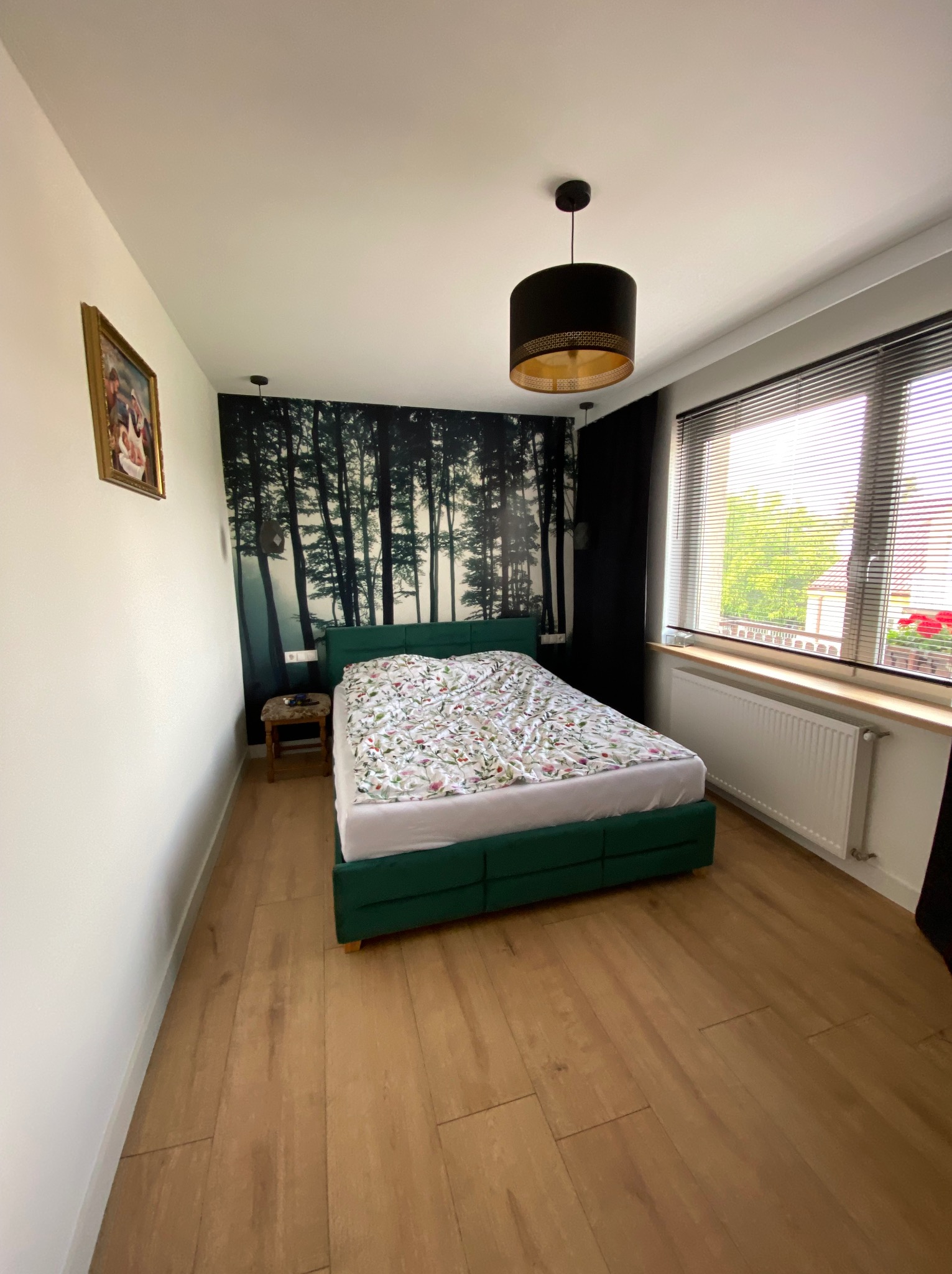 Sypialnia z drewnianą podłogą, zielonym łóżkiem i fototapetą z motywem lasu, oświetlona lampą z czarnym abażurem i złotym wnętrzem.