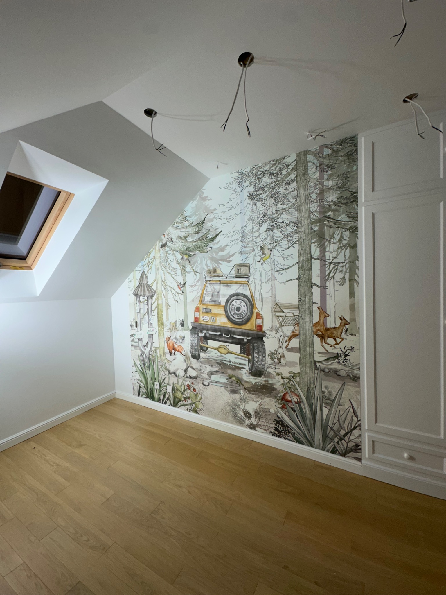 Pokój na poddaszu z drewnianą podłogą i fototapetą przedstawiającą leśny krajobraz z samochodem terenowym, widoczne niedokończone instalacje elektryczne na suficie.