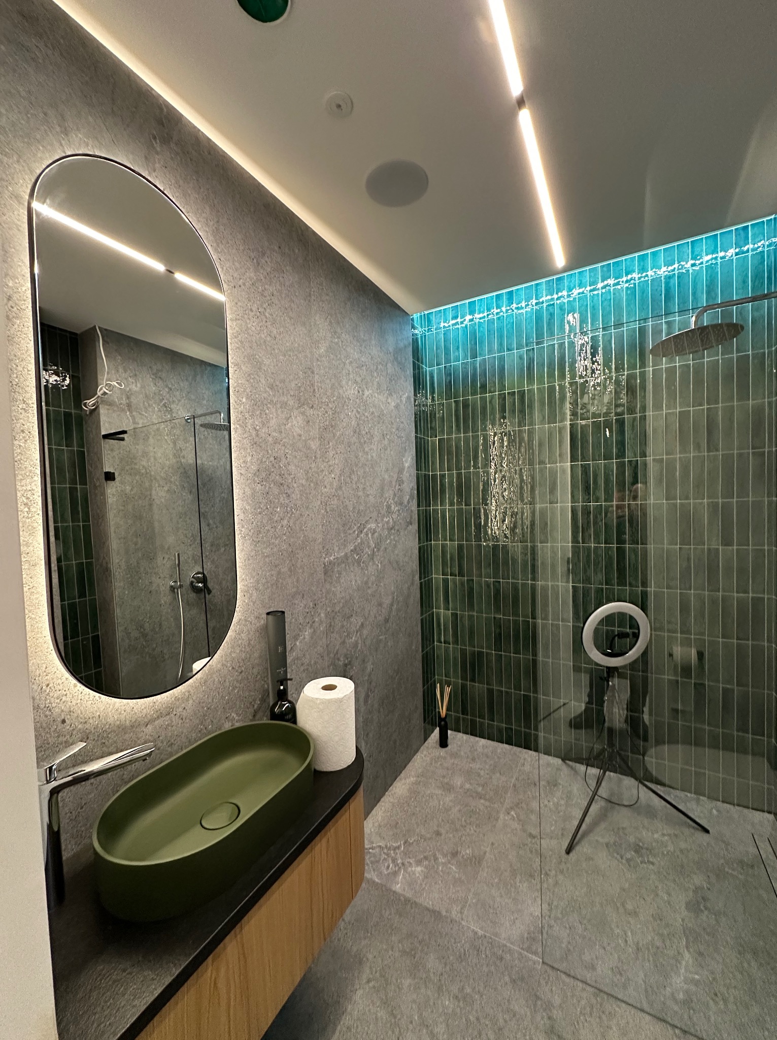 Nowoczesna łazienka z zielonymi płytkami prysznicowymi podświetlonymi na niebiesko, owalną umywalką w kolorze oliwkowym i lustrem z podświetleniem LED. Widoczny również ring do nagrywania filmów.