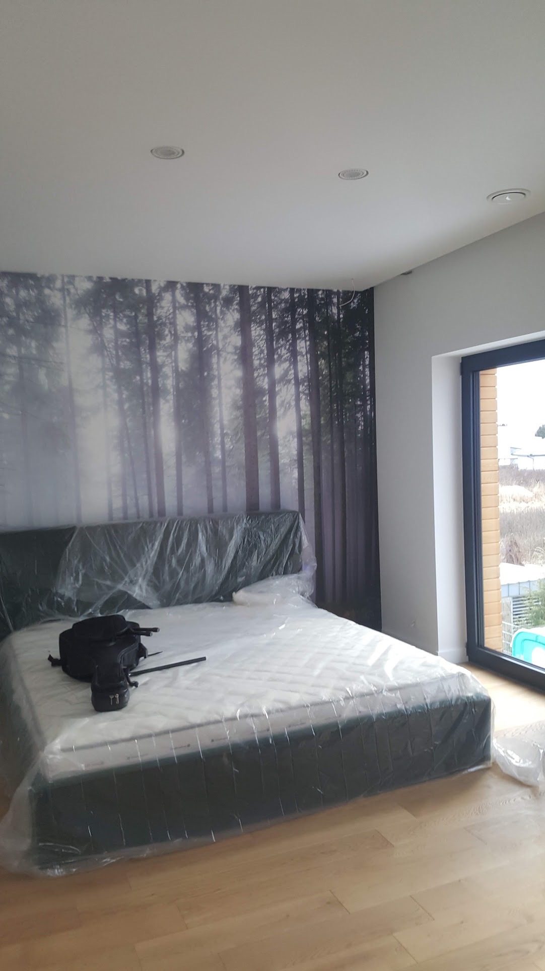 Nowe łóżko z materacem zabezpieczone folią ochronną w pokoju z fototapetą imitującą las, widoczne drewniane panele podłogowe i czarna torba.