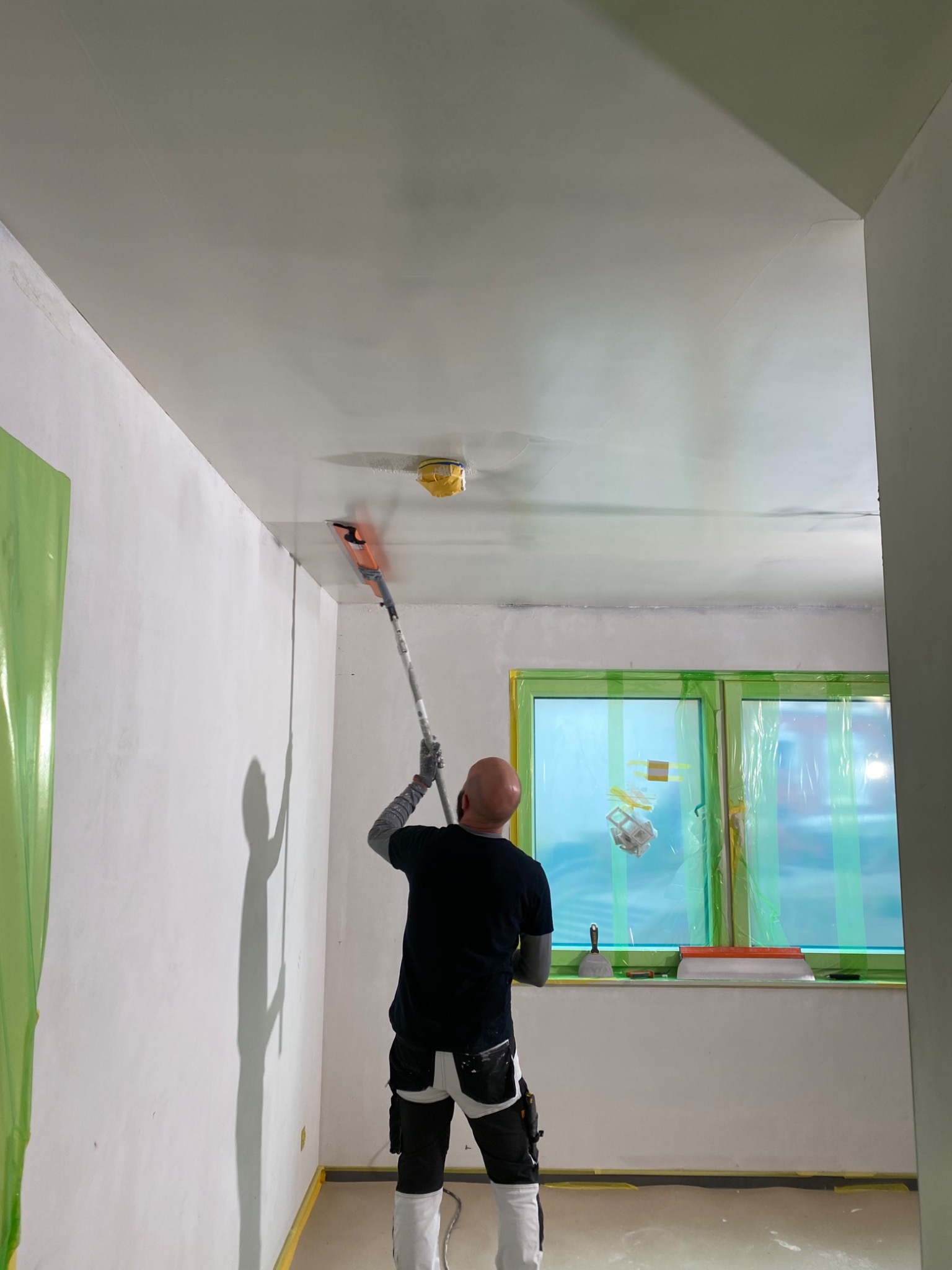 Mężczyzna w czarnej koszulce i roboczych spodniach, z widocznymi narzędziami, wygładza sufit za pomocą szpachli na długim kiju; okno zabezpieczone folią, widoczne narzędzia na parapecie.