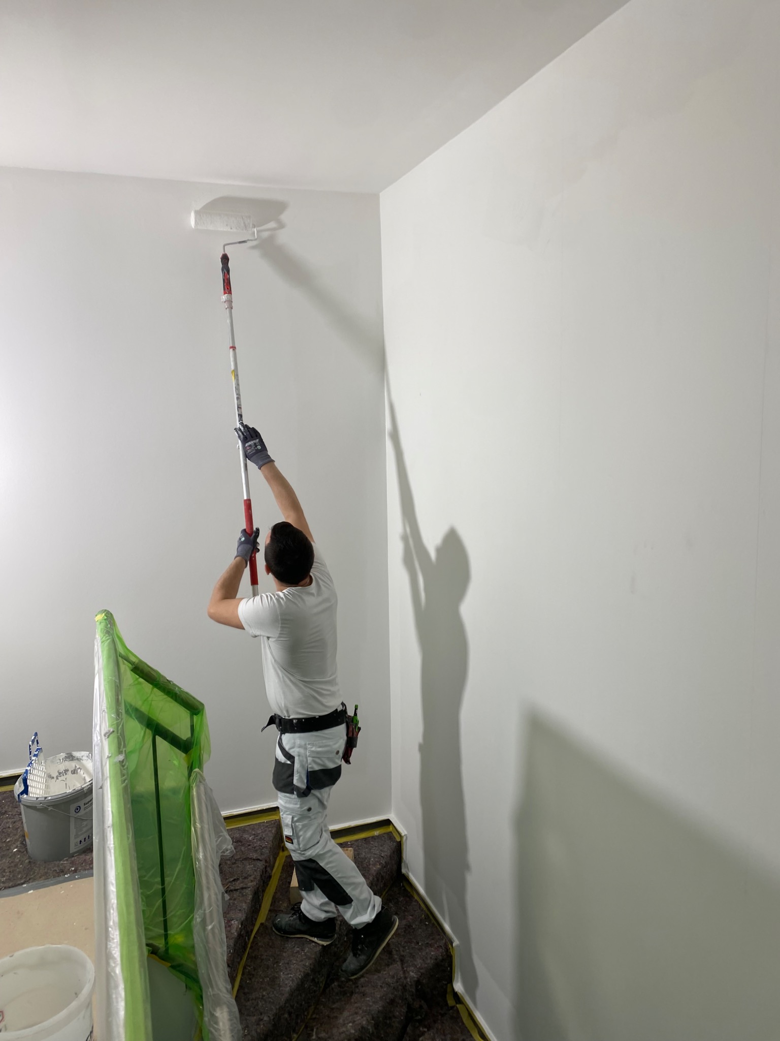 Mężczyzna w roboczym ubraniu maluje białą ścianę wałkiem na długim teleskopowym uchwycie, widoczne zabezpieczone schody i wiadro z farbą.