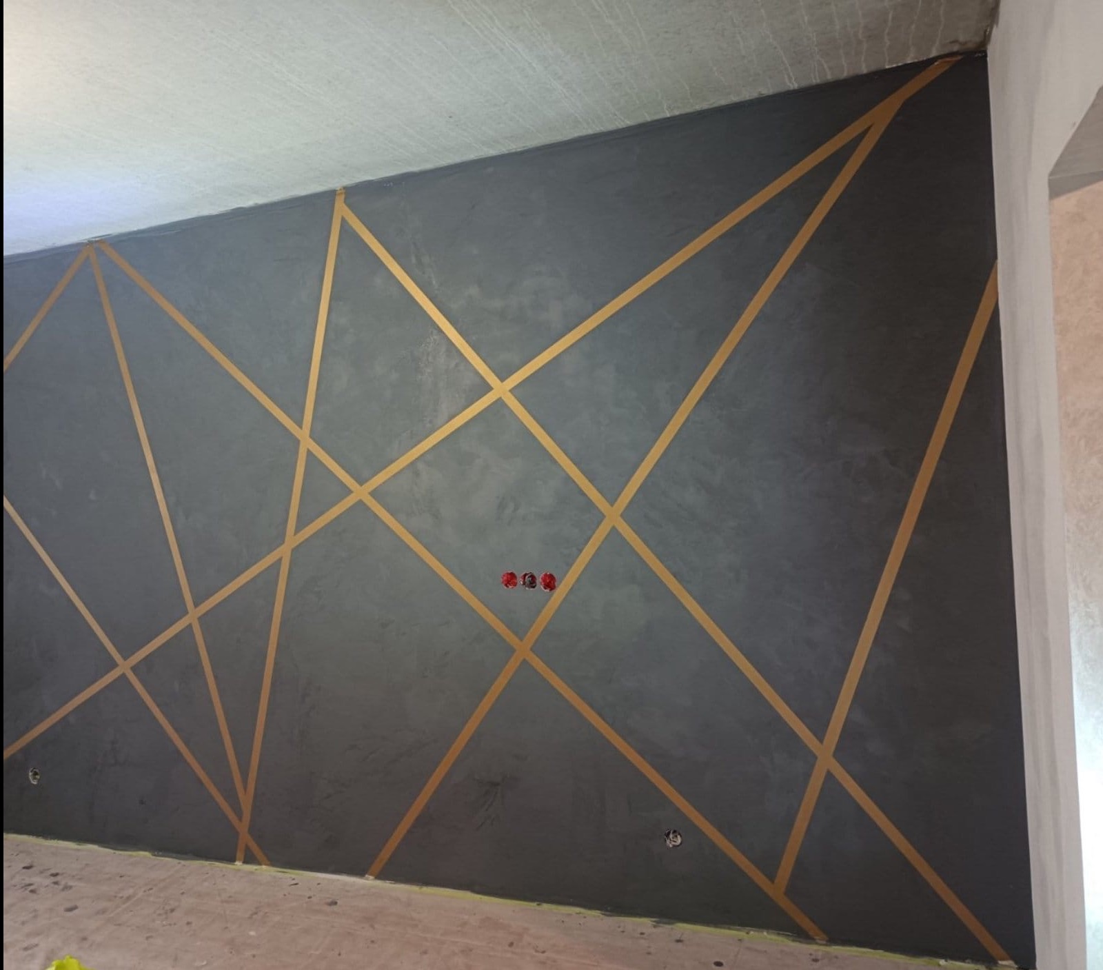 Szara ściana z dekoracyjną strukturą i złotymi geometrycznymi pasami, widoczne gniazdka elektryczne.