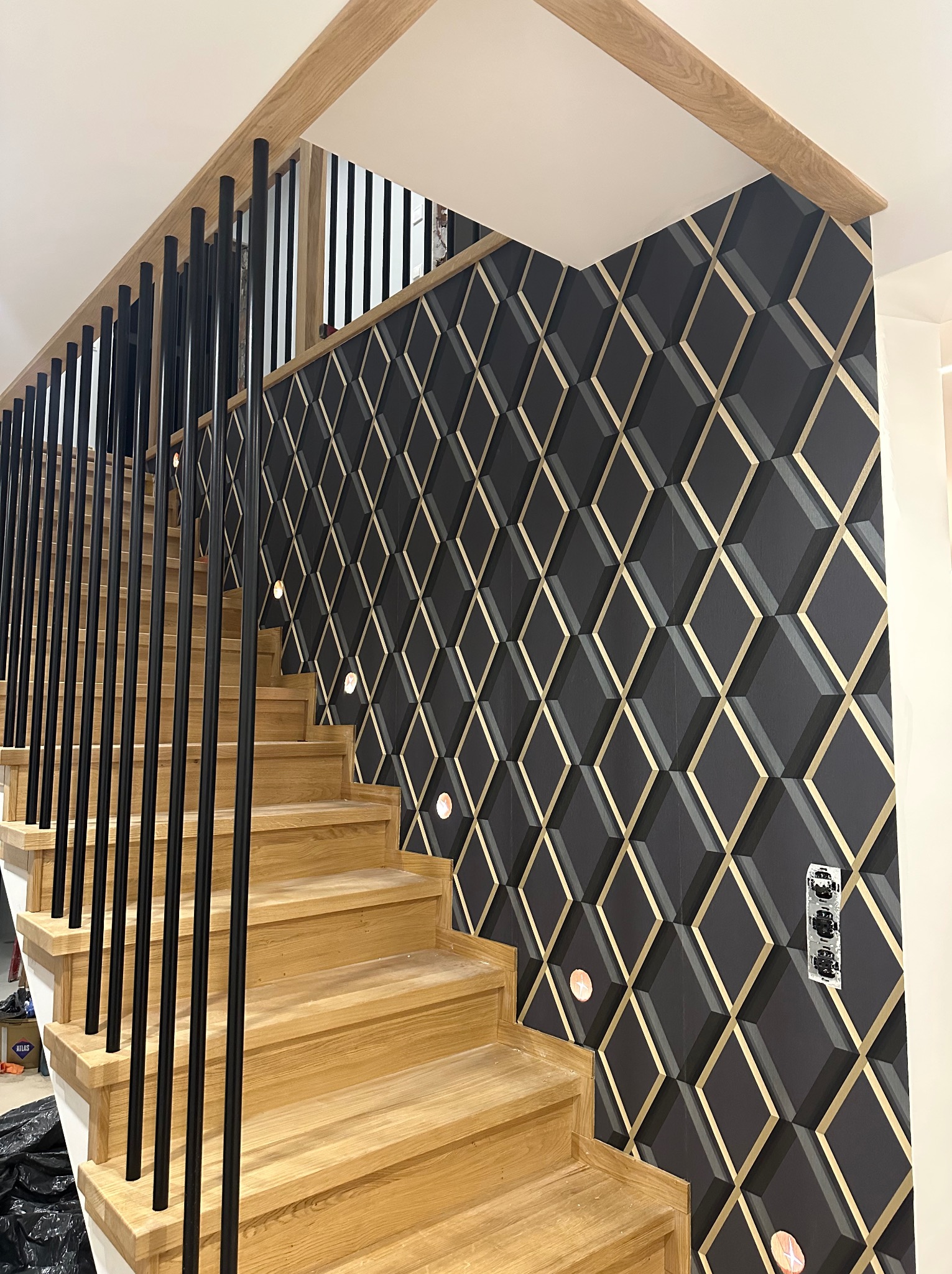 Nowoczesne schody drewniane z czarną balustradą i ścianą pokrytą tapetą 3D w geometryczne wzory z elementami złota, oświetlenie punktowe w ścianie.