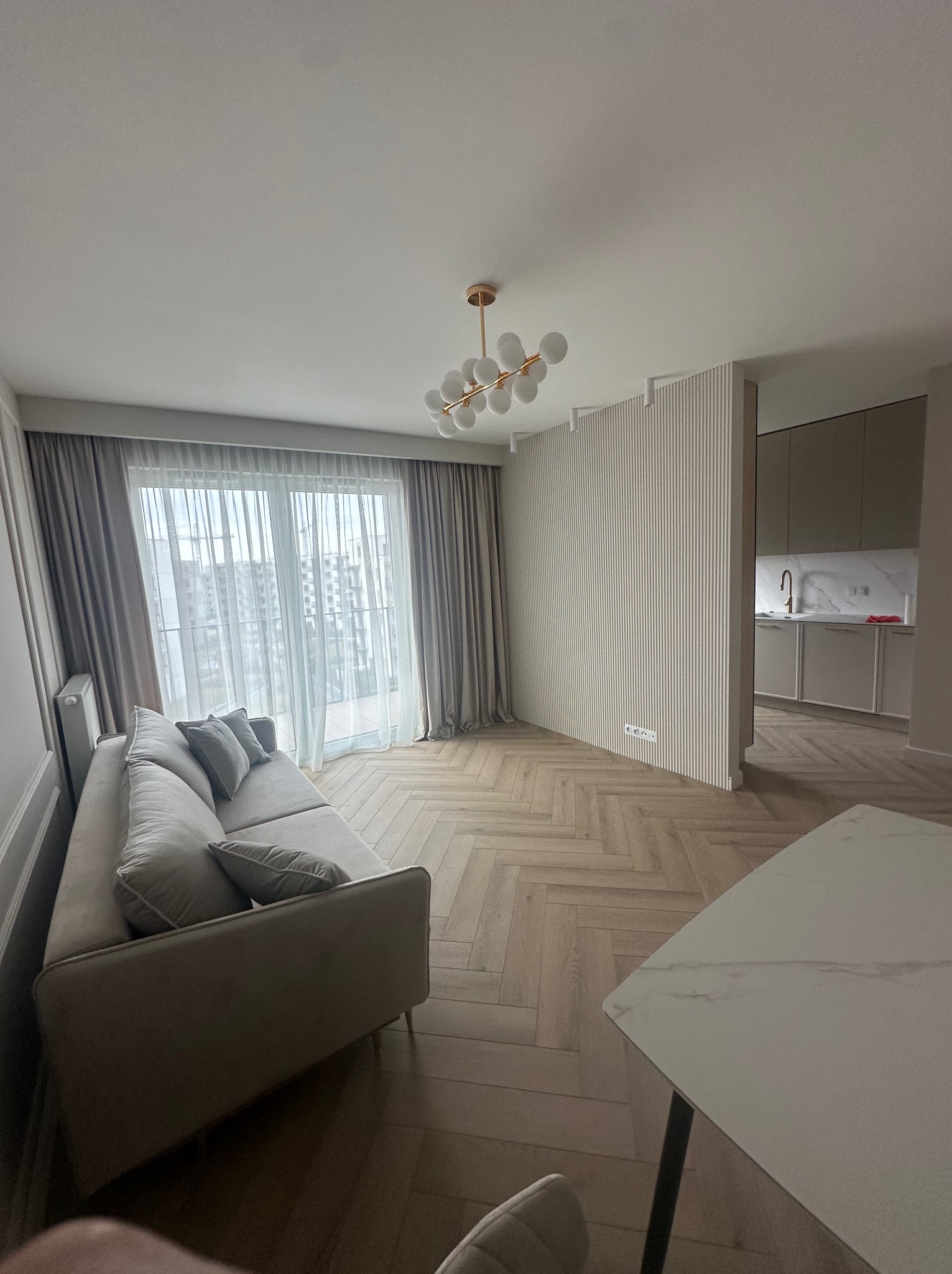 Jasne wnętrze salonu z drewnianą podłogą ułożoną w jodełkę, szarą sofą z poduszkami, białymi zasłonami i widokiem na kuchnię z marmurowym blatem.