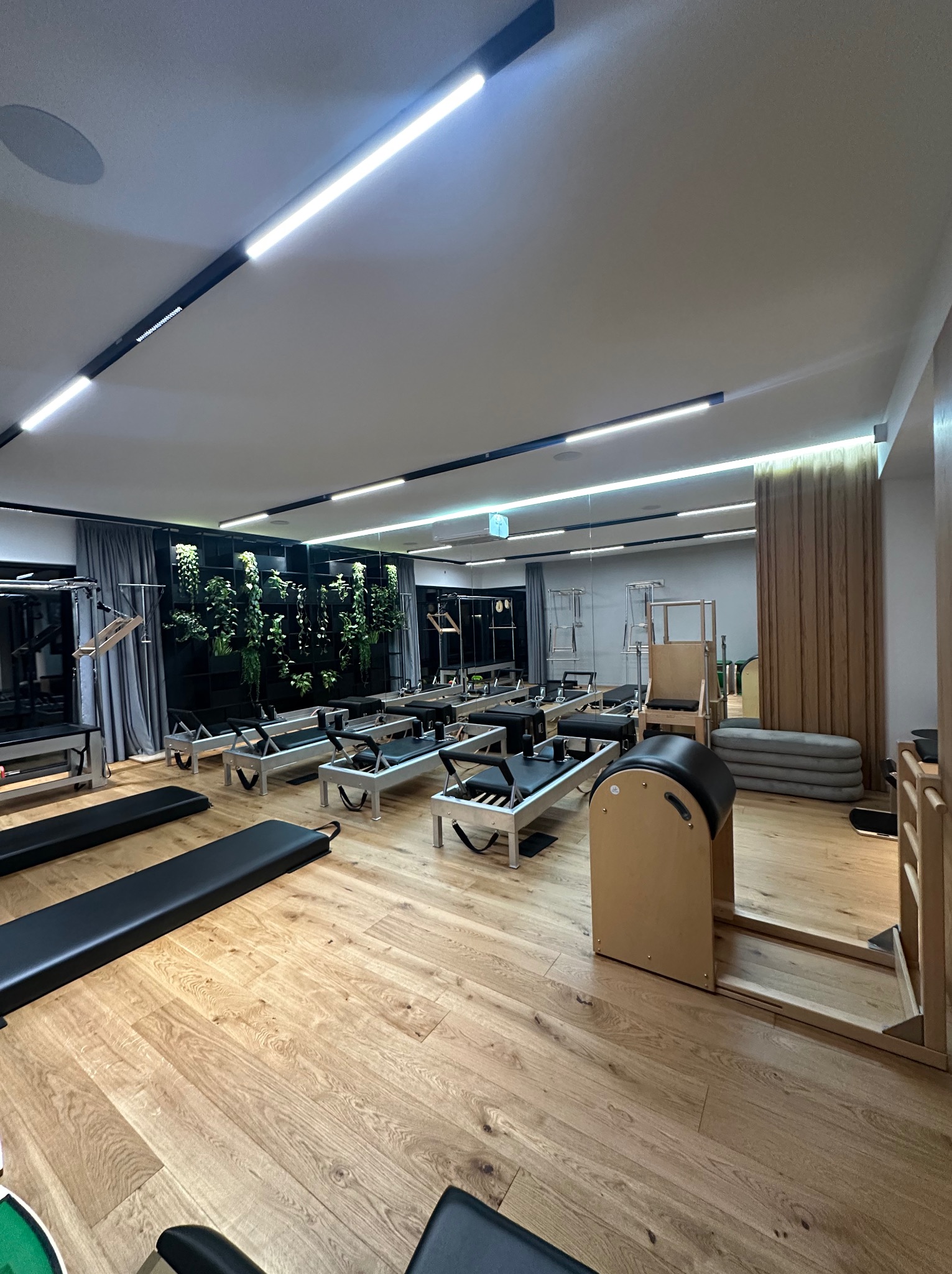 Jasna sala do ćwiczeń z drewnianą podłogą, wyposażona w sprzęt do ćwiczeń typu reformer i ścianę z roślinami.