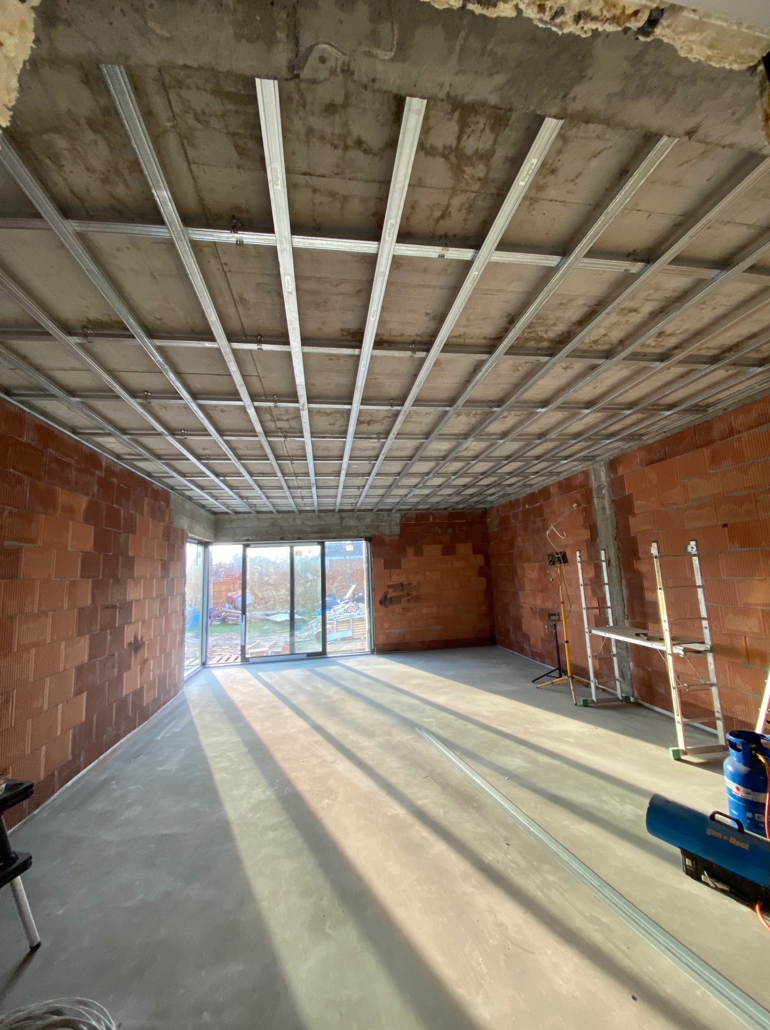 Surowy stan wnętrza z widoczną konstrukcją metalową pod sufitem podwieszanym, ściany z czerwonej cegły, betonowa podłoga, drabina i nagrzewnica. Przygotowania do prac wykończeniowych.