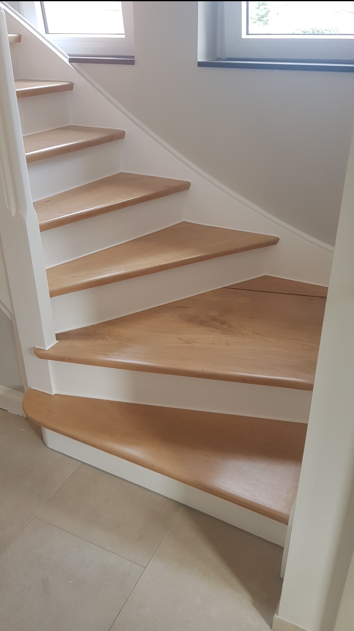 Jasne drewniane schody z białymi podstopnicami i poręczą, widok z dołu, neutralne płytki podłogowe, naturalne światło z okna w tle.