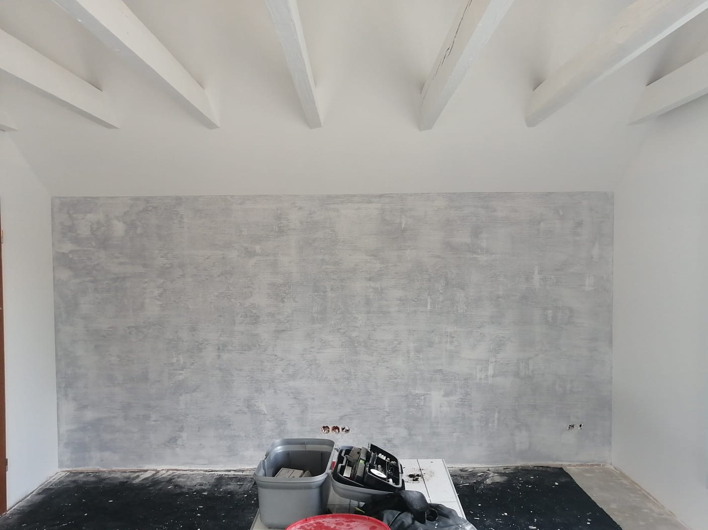 Ściana w pokoju po nałożeniu dekoracyjnej szarej struktury, widoczne białe belki stropowe, przygotowane narzędzia i zabezpieczona podłoga.