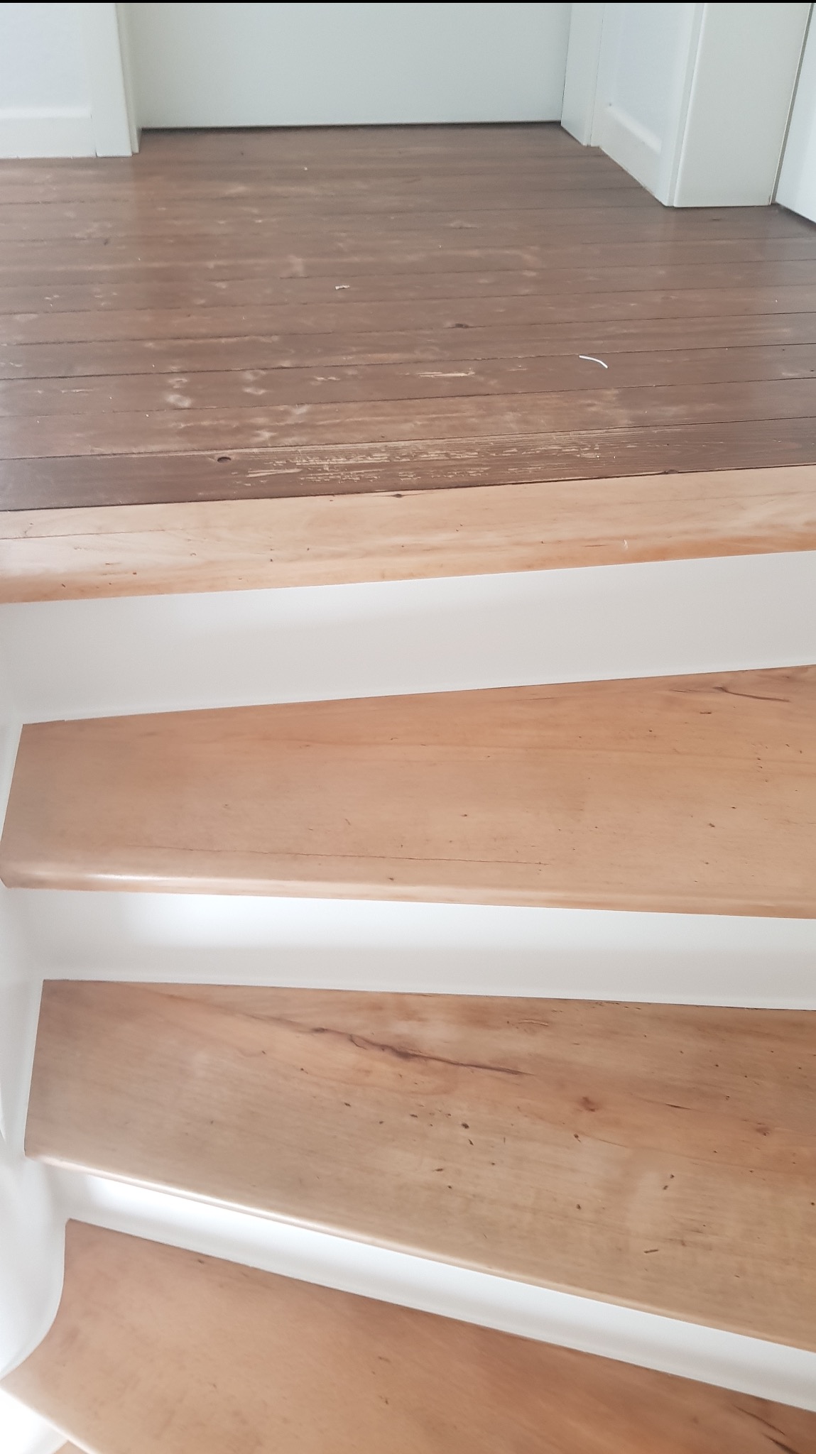 Schody drewniane z jasnego drewna, białe podstopnice, widoczny fragment podłogi z ciemniejszego drewna z widocznymi śladami zużycia.