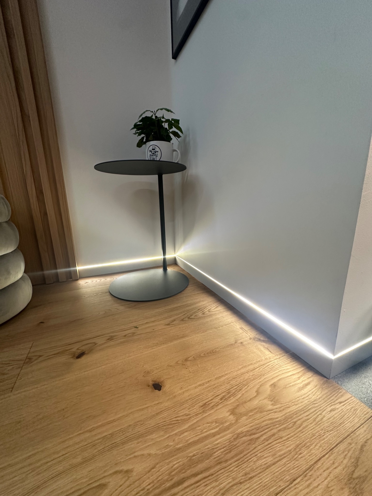 Nowoczesne wnętrze z drewnianą podłogą, listwą przypodłogową z podświetleniem LED, minimalistycznym stolikiem z rośliną w kubku i elementem dekoracyjnym z drewnianymi listwami na ścianie.