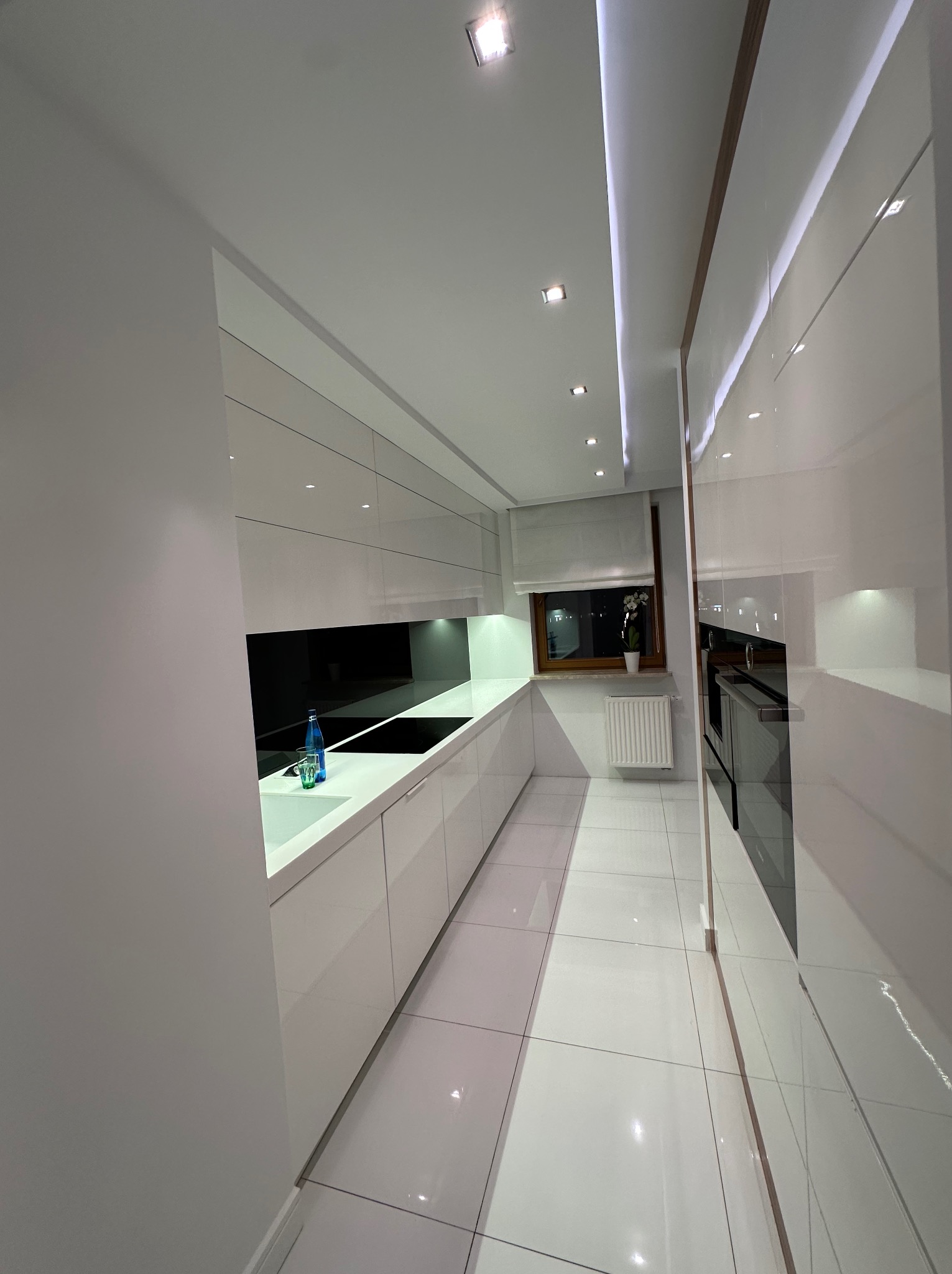 Wnętrze nowoczesnej kuchni z białymi, lakierowanymi szafkami, czarnym panelem nad blatem i ceramiczną podłogą. Na blacie stoi szklana butelka z wodą i szklanka. Widoczne oświetlenie sufitowe i okno...