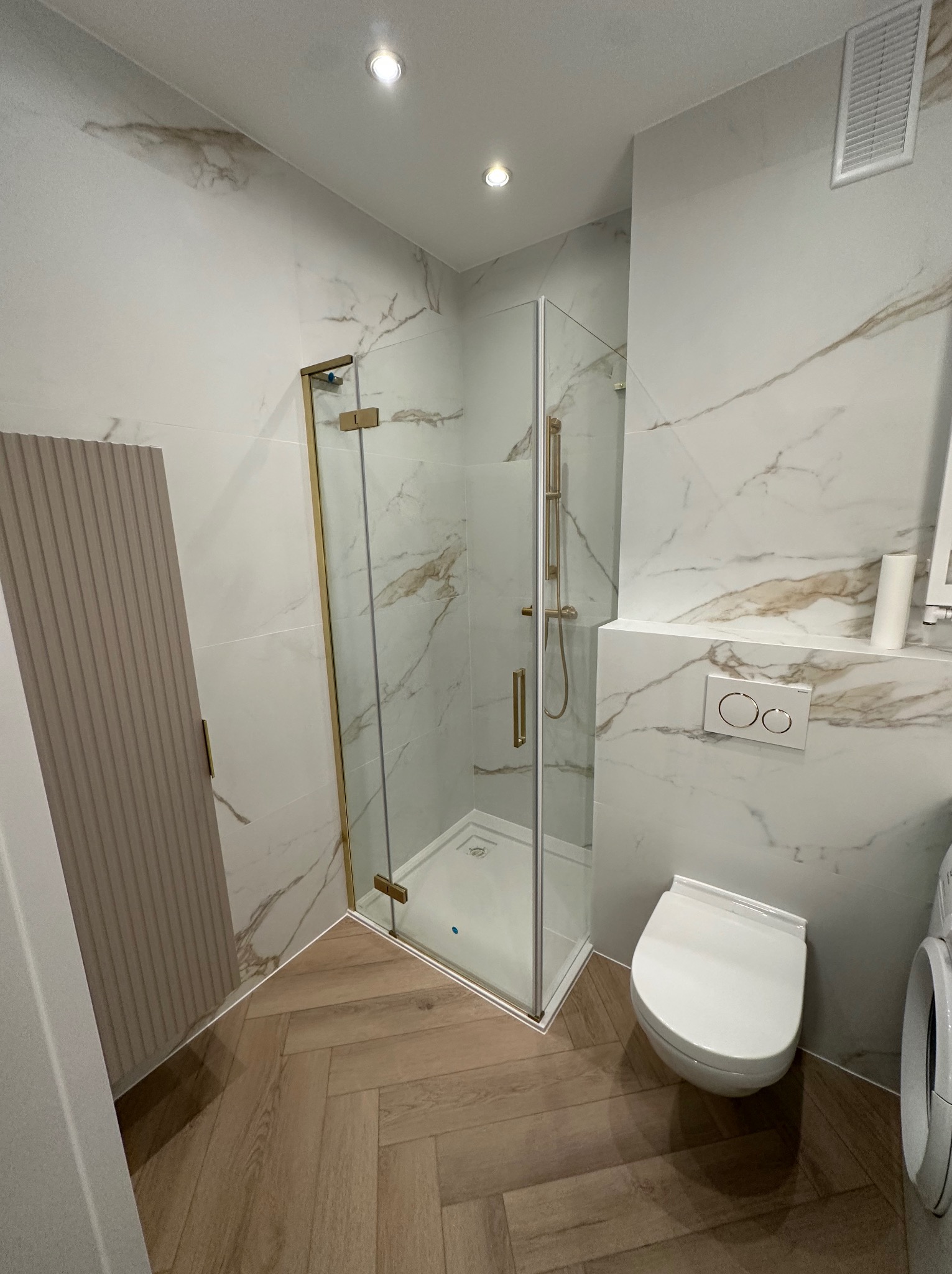 Nowoczesna łazienka z kabiną prysznicową w złotej ramie, toaletą podwieszaną i płytkami imitującymi marmur, podłoga z paneli ułożonych w jodełkę.