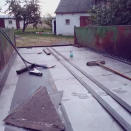 Wnętrze metalowej konstrukcji w trakcie spawania, widoczne narzędzia spawalnicze, szczotka, puszka z preparatem, resztki metalu i ślady spawania, w tle budynki gospodarcze.