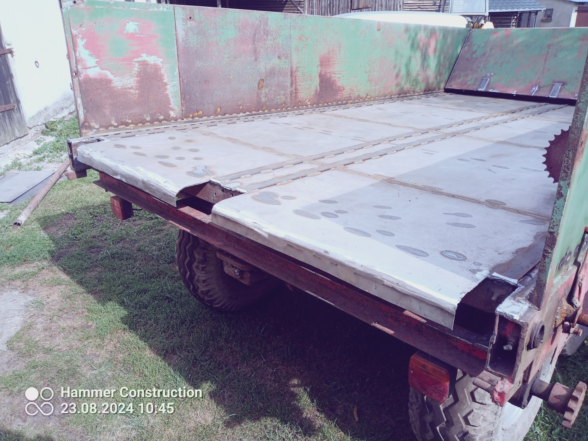 Przyczepa rolnicza w trakcie naprawy: nowa, spawana podłoga z blachy na tle starej, zielonej ramy z widoczną korozją i śladami spawania. Widoczne opony i fragment trawnika.