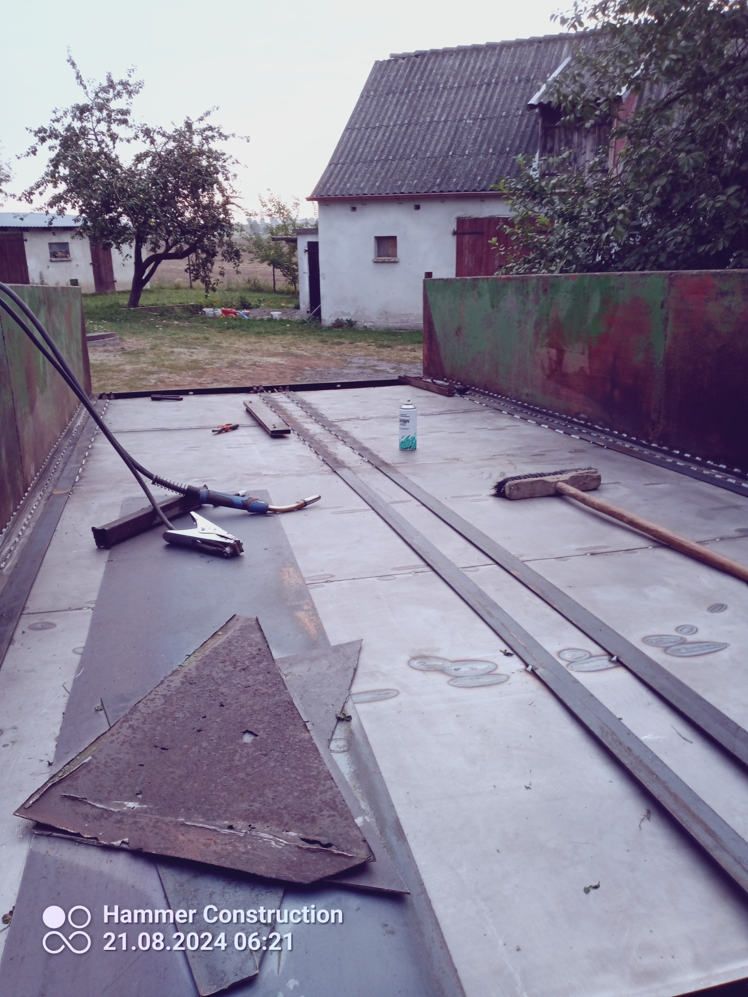 Wnętrze metalowej konstrukcji w trakcie spawania, widoczne narzędzia spawalnicze, szczotka, puszka z preparatem, resztki metalu i ślady spawania, w tle budynki gospodarcze.