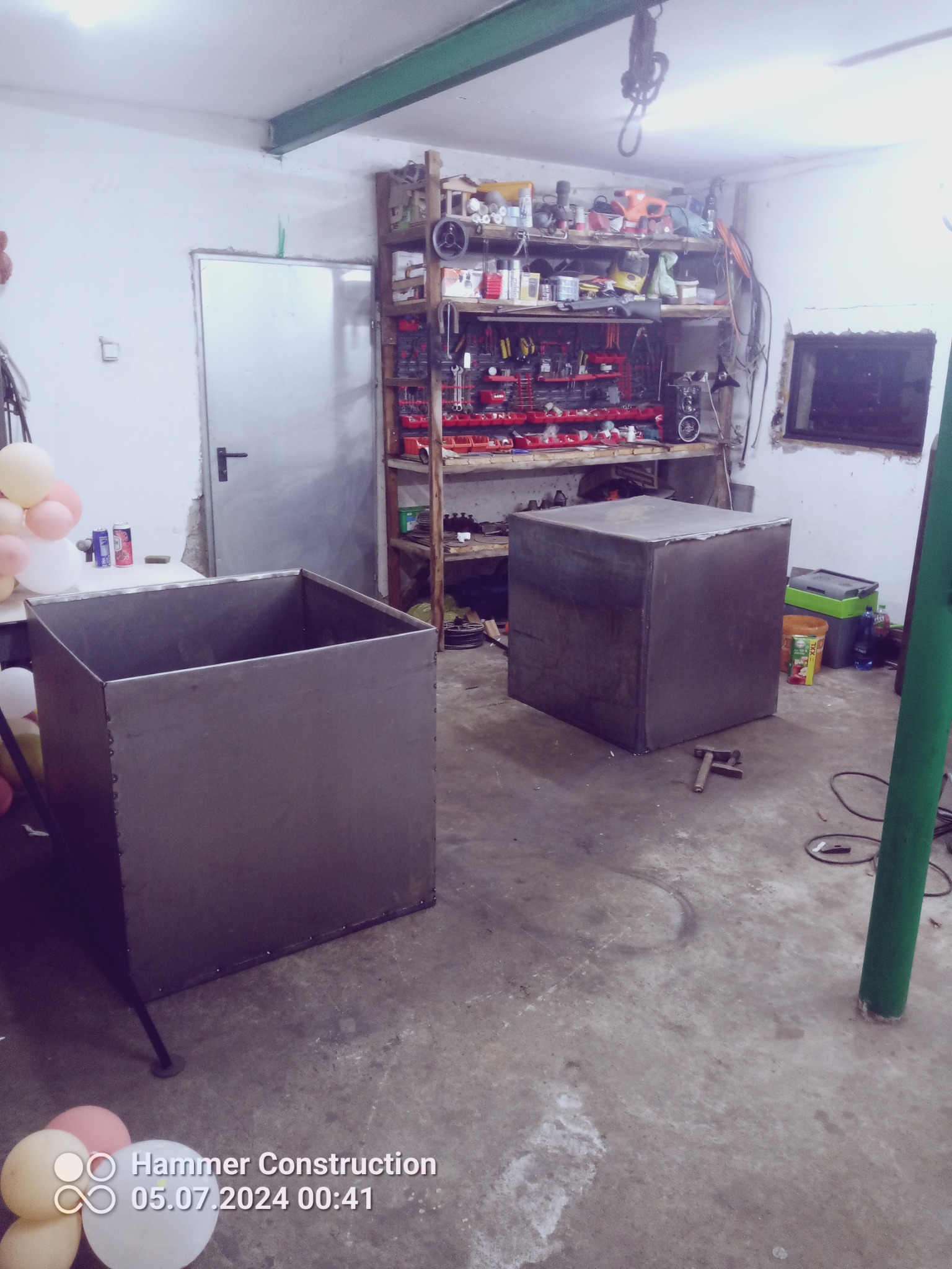 Dwa metalowe, surowe, kwadratowe kontenery z widocznymi śladami spawania stoją w warsztacie. Jeden kontener ma nogi, drugi stoi bezpośrednio na podłodze. W tle widoczne narzędzia na półkach i drzwi.