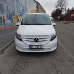 Biały Mercedes Vito zaparkowany na chodniku z kostki brukowej, na tle budynku z muralem przedstawiającym sceny historyczne.