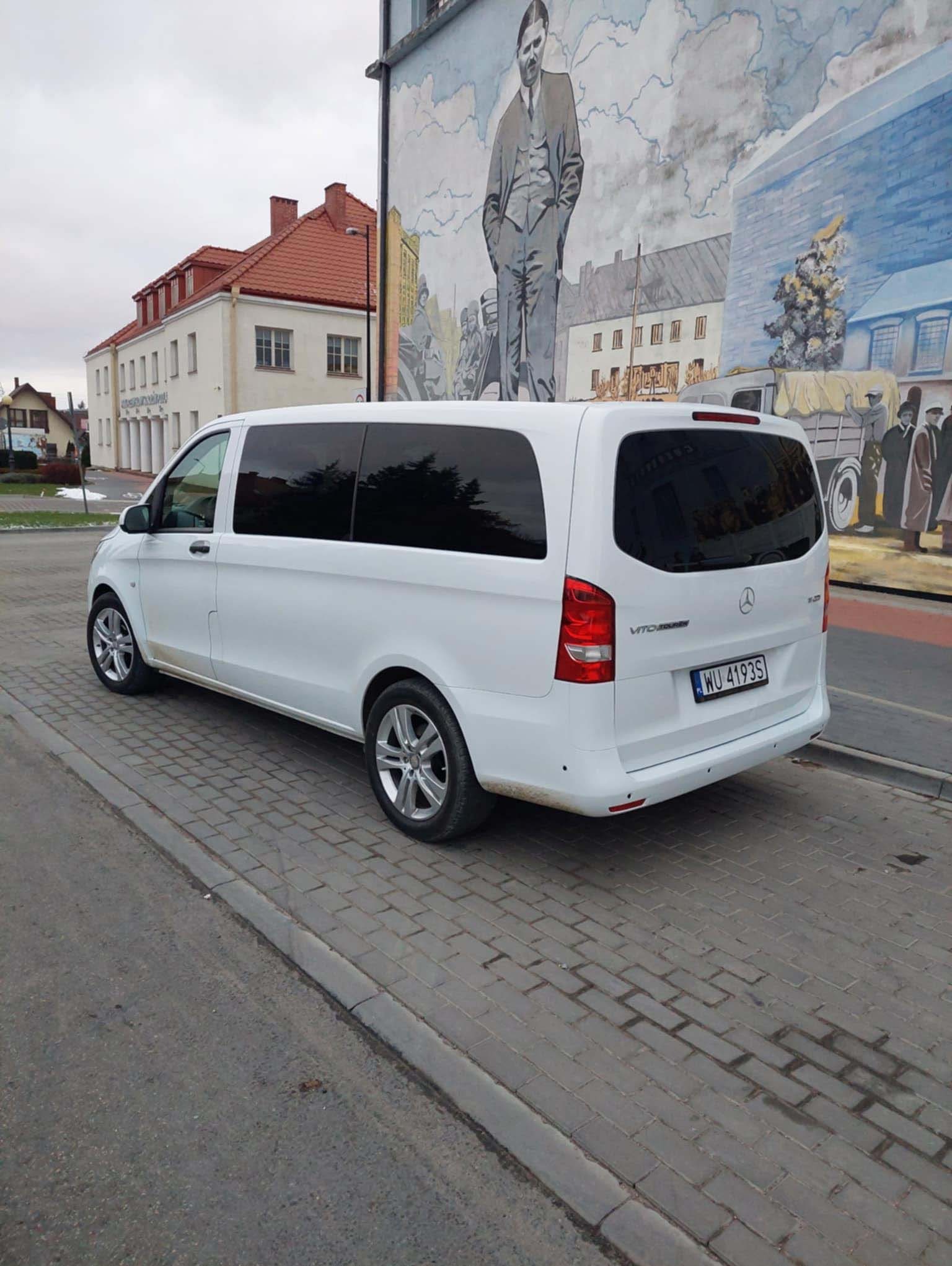 Biały Mercedes Vito z przyciemnianymi szybami zaparkowany na chodniku z kostki brukowej, na tle budynku z czerwonym dachem i muralu przedstawiającego postać w garniturze.