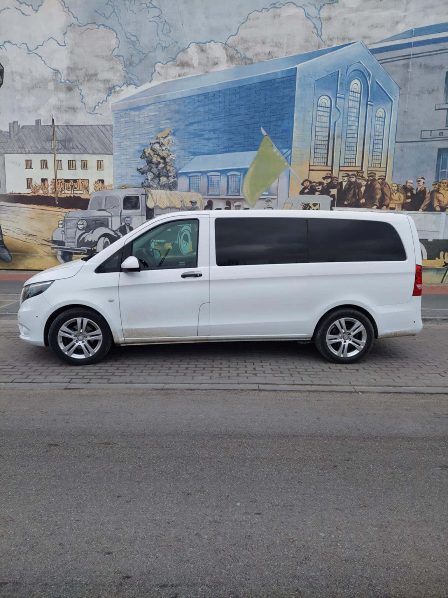 Biały bus Mercedes Vito zaparkowany na chodniku przed ścianą z muralem przedstawiającym sceny historyczne.