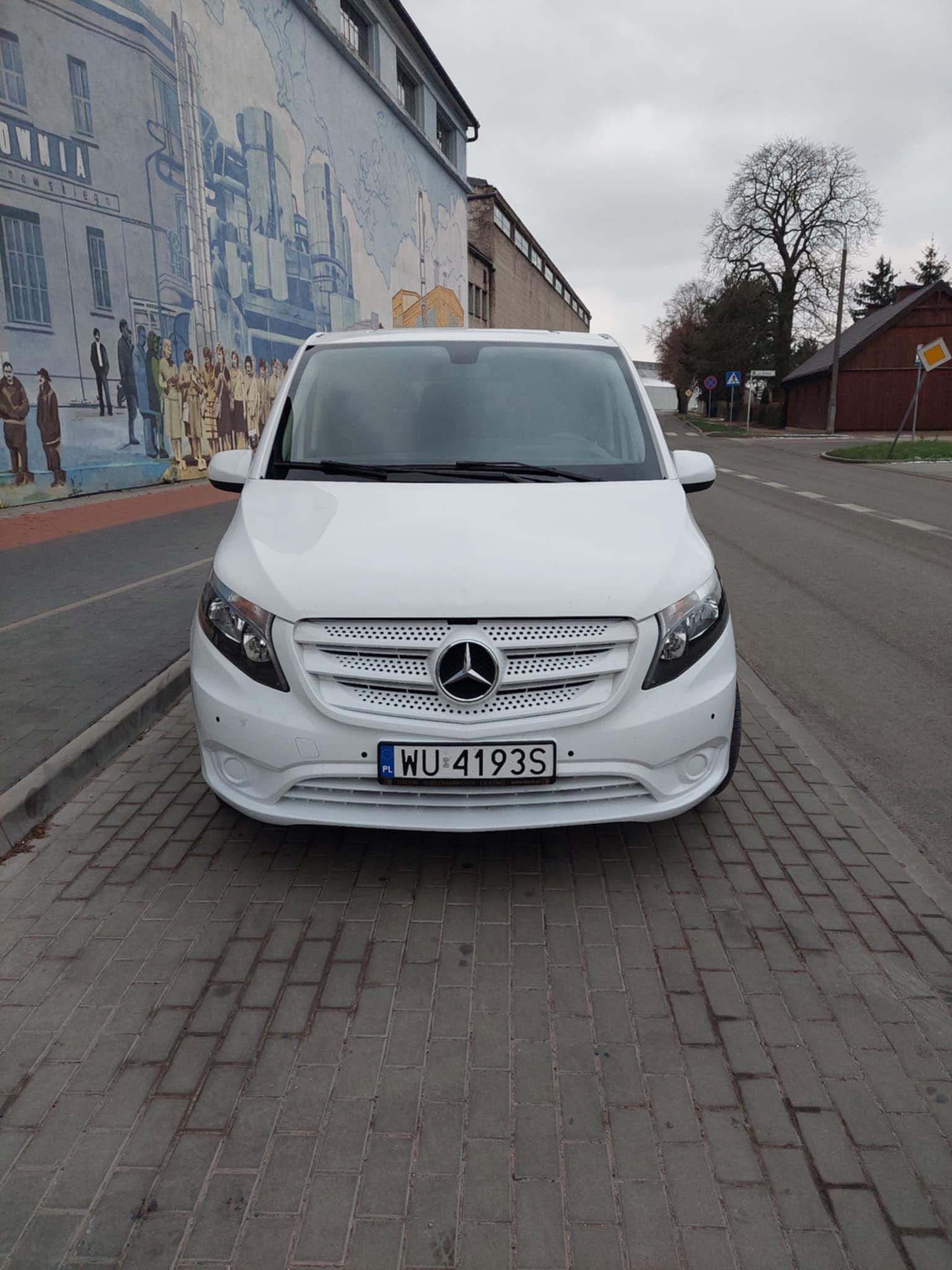 Biały Mercedes Vito zaparkowany na chodniku z kostki brukowej, na tle budynku z muralem przedstawiającym sceny historyczne.