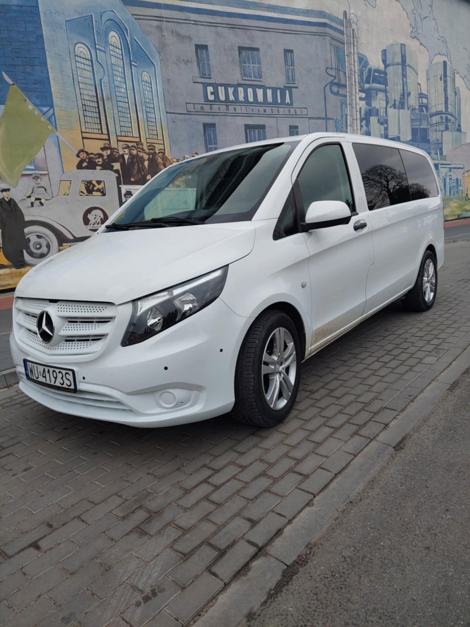 Biały Mercedes Vito zaparkowany na chodniku z kostki brukowej na tle muralu przedstawiającego budynek Cukrowni i ludzi.