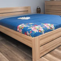 Łóżko z jasnego drewna z niebieską narzutą w kwiaty, książką i okularami na wierzchu, obok szafka nocna z małym drzewkiem bonsai, w tle komoda i okno z firanką.