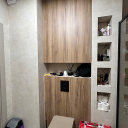 LUK-MEB Meble na wymiar - Zabudowa toalety z szafkami z drewnopodobnej płyty i wnękami w ścianie. U dołu widoczny sedes i kosz na śmieci. Na podłodze karton z drukarką.