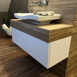 LUK-MEB Meble na wymiar - Nowoczesna umywalka nablatowa z szafką w łazience, drewniany blat i białe fronty, w tle ściana z drewnopodobnych płytek i roślina w ozdobnej donicy.