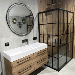 LUK-MEB Meble na wymiar - Nowoczesna łazienka z drewnianą zabudową umywalki z białym blatem, okrągłym lustrem i kabiną prysznicową w stylu loft na tle szarych i drewnianych płytek.