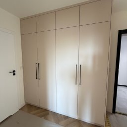 LUK-MEB Meble na wymiar - Beżowa szafa wnękowa z czarnymi uchwytami, dopasowana do wnętrza z parkietem w jodełkę. Minimalistyczny design, widok na fragment drzwi i sufit z lampą.
