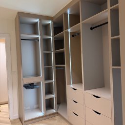 LUK-MEB Meble na wymiar - Jasna, narożna szafa wnękowa z oświetleniem LED, widoczne półki, drążki i szuflady z charakterystycznymi uchwytami. Podłoga z parkietu w jodełkę.