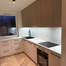 LUK-MEB Meble na wymiar - Nowoczesna kuchnia z beżowymi frontami, czarnymi uchwytami i armaturą. Blat imitujący marmur, podświetlenie LED i parkiet w jodełkę tworzą elegancki wystrój.