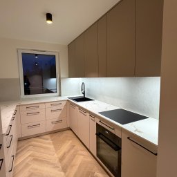 LUK-MEB Meble na wymiar - Nowoczesna kuchnia w kształcie litery U z beżowymi frontami, czarnymi uchwytami i blatem imitującym marmur. Na blacie czarna płyta indukcyjna i zlew. Parkiet ułożony w jodełkę.