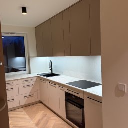 LUK-MEB Meble na wymiar - Nowoczesna kuchnia na wymiar z beżowymi frontami, czarną armaturą i blatem imitującym marmur. Widoczna płyta indukcyjna i piekarnik. Parkiet ułożony w jodełkę.